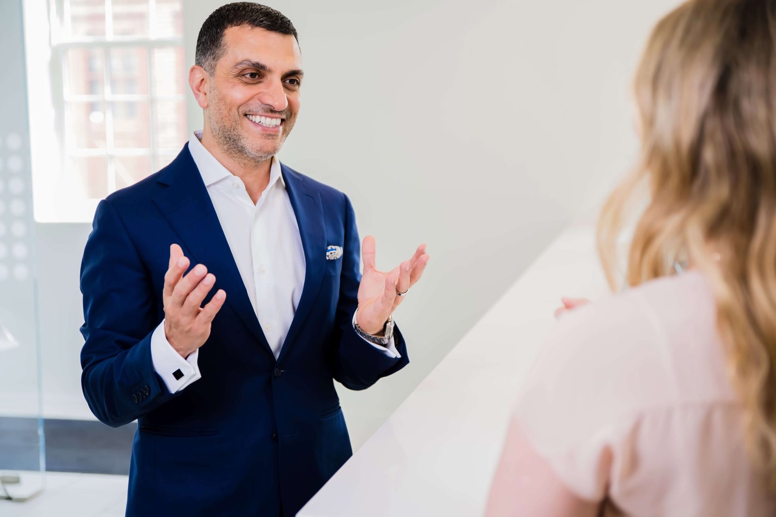 A male clinician in a smart blue suit engaging in a positive conversation with a female patient in a clinic setting. The clinician uses expressive hand gestures, suggesting discussion about patient care or treatment options. The professional and inviting atmosphere highlights effective patient communication, relevant to themes of consistent clinical care and recovery strategies discussed in the blog article.