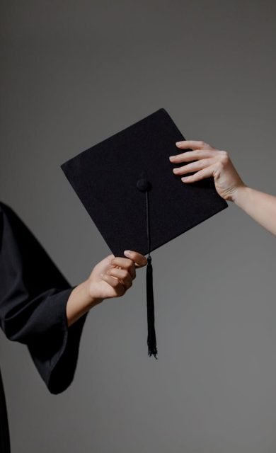 Two people exchanging a graduation cap on a grey background, symbolizing achievement.