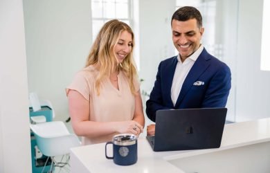 Two professionals in a modern clinic setting collaborate and review something on a laptop, highlighting teamwork, communication, and training often found in high-performing chiropractic teams. The scene is bright, wide, and inviting, conveying operational excellence and a seamless patient experience.